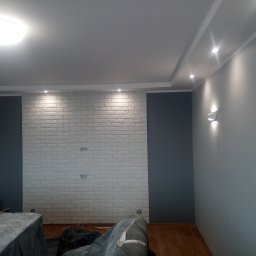 krzysztof kaleński - Pokój po remoncie: ściana z białej cegły dekoracyjnej, fragment szarej ściany, sufit z oświetleniem punktowym i centralnym, zabezpieczone meble folią malarską.