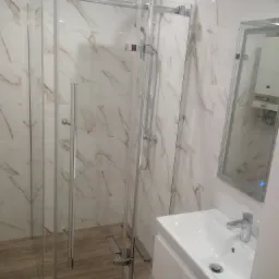 Nowoczesna łazienka z kabiną prysznicową typu walk-in, białą umywalką z szafką i lustrem w ramie, wykończona płytkami imitującymi marmur.