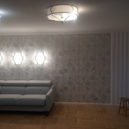 krzysztof kaleński - Szary narożnik z podnoszonymi zagłówkami na tle ściany z efektem betonu, oświetlonej trzema kinkietami i dwoma lampami sufitowymi, na parkiecie, z widocznym stołkiem barowym w rogu.