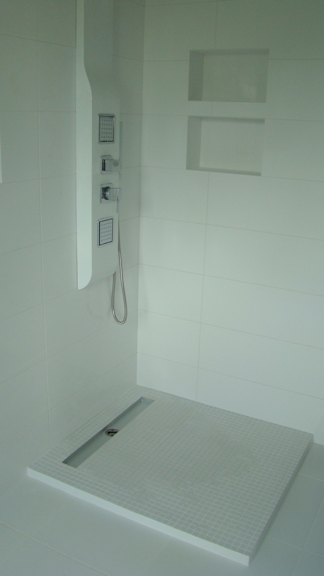 Biała kabina prysznicowa z odpływem liniowym i panelem natryskowym z hydromasażem, wykończona mozaiką i wnękami w ścianie.