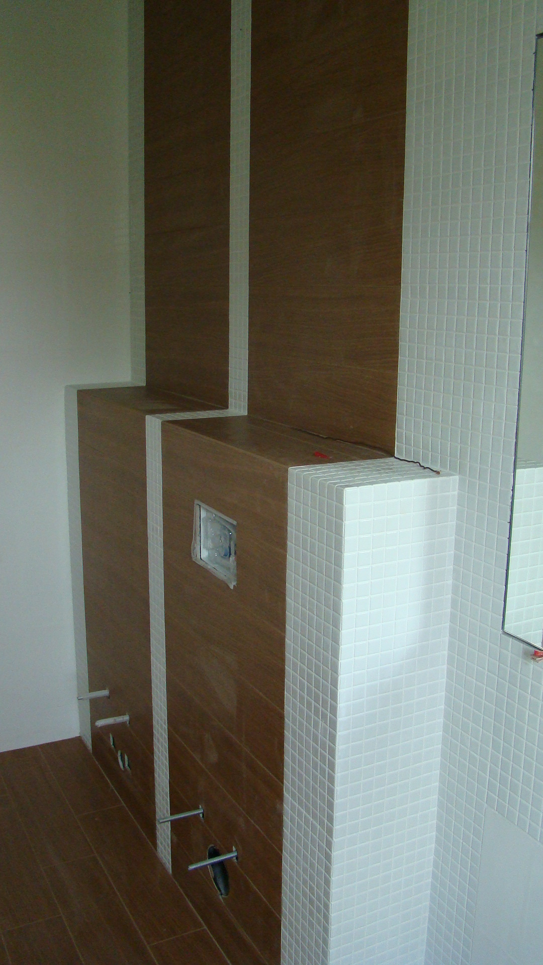 Wnęka zabudowy stelaża WC z drewnopodobnymi panelami i białymi mozaikami, widoczne wystające elementy montażowe, etap prac wykończeniowych.