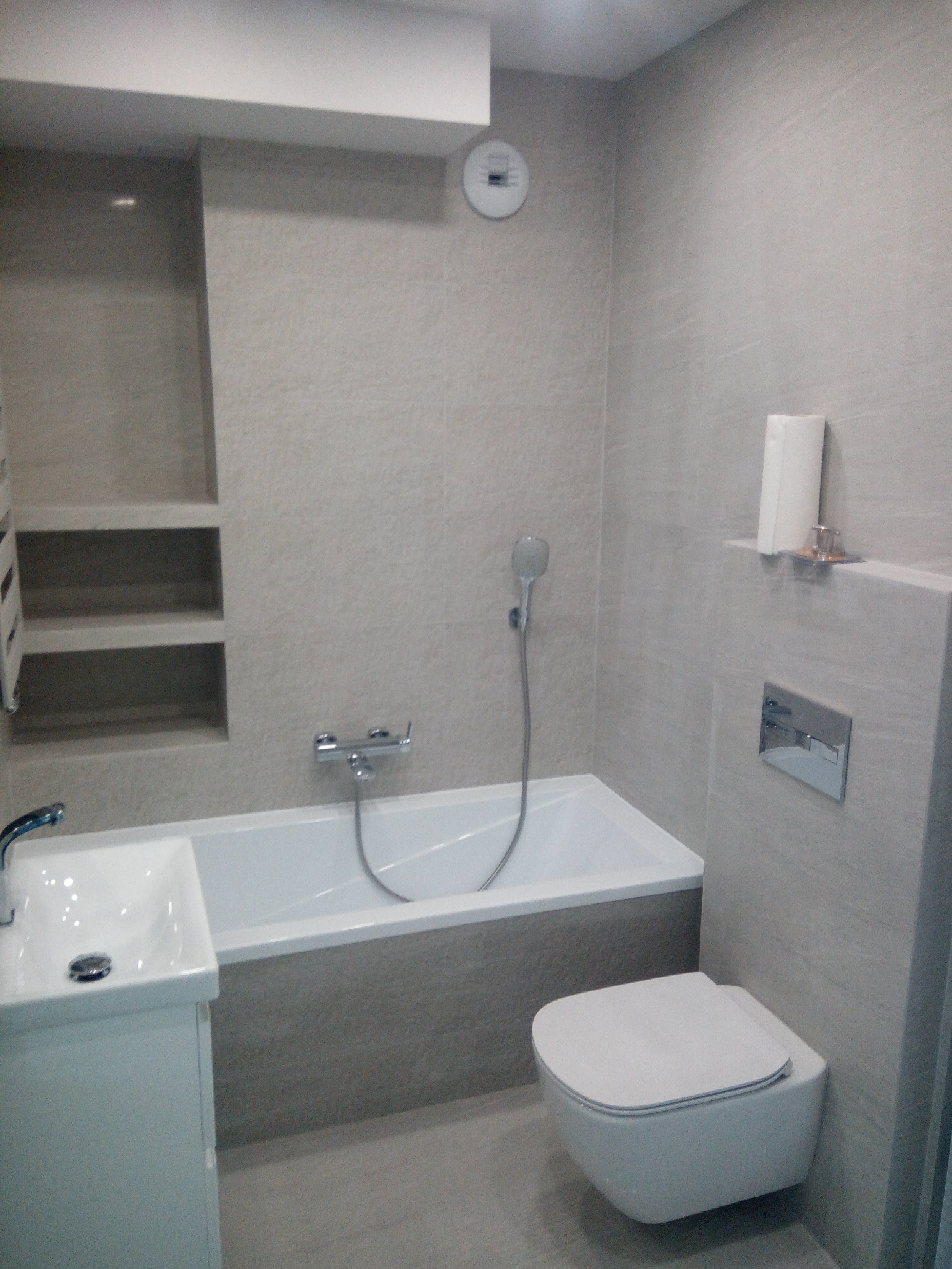 Nowoczesna, jasnoszara łazienka w Kielcach po remoncie, z wiszącą toaletą, wanną z prysznicem i umywalką z szafką, utrzymana w minimalistycznym stylu.