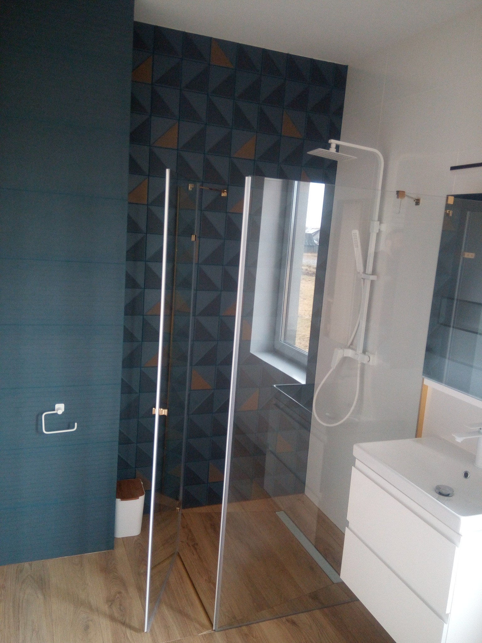 Nowoczesna łazienka z kabiną prysznicową typu walk-in, wykończona płytkami w odcieniach granatu i drewna, z widocznym odpływem liniowym w podłodze i minimalistyczną białą umywalką z szafką.