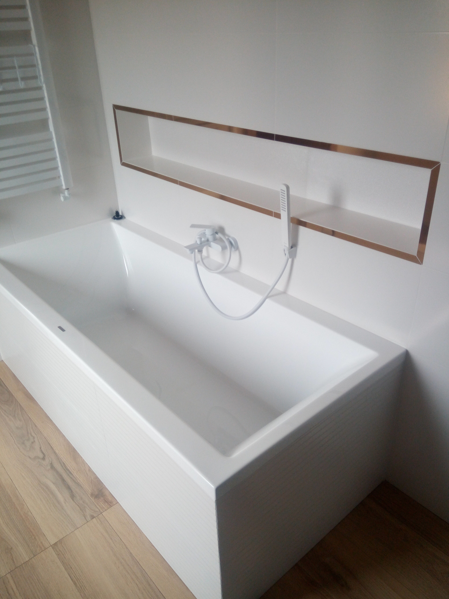 Biała, prostokątna wanna z baterią i słuchawką prysznicową, wbudowana w ścianę półka z dekoracyjną, brązową ramką, jasne płytki na ścianach i podłodze imitującej drewno.