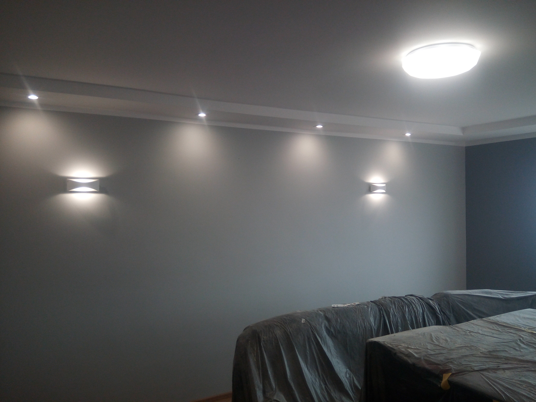 Świeżo pomalowana szara ściana z zamontowanymi kinkietami i oświetleniem punktowym w suficie, meble zabezpieczone folią ochronną.