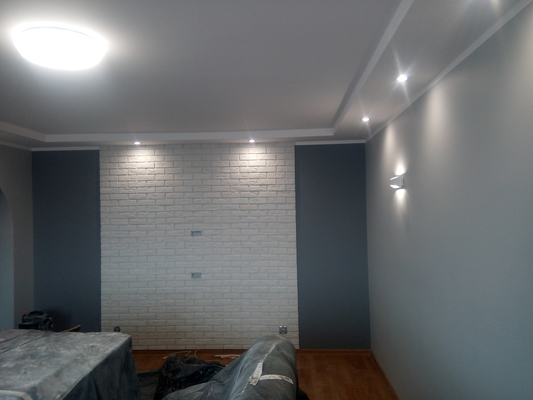Pokój po remoncie: ściana z białej cegły dekoracyjnej, fragment szarej ściany, sufit z oświetleniem punktowym i centralnym, zabezpieczone meble folią malarską.