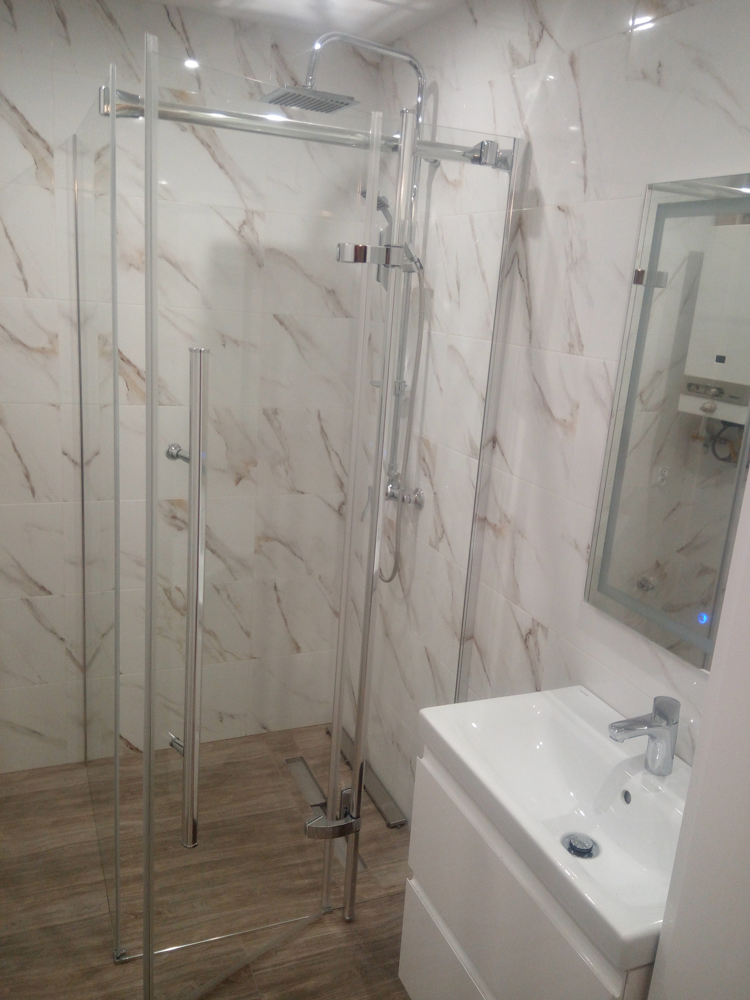 Nowoczesna łazienka z kabiną prysznicową typu walk-in, białą umywalką z szafką i lustrem w ramie, wykończona płytkami imitującymi marmur.