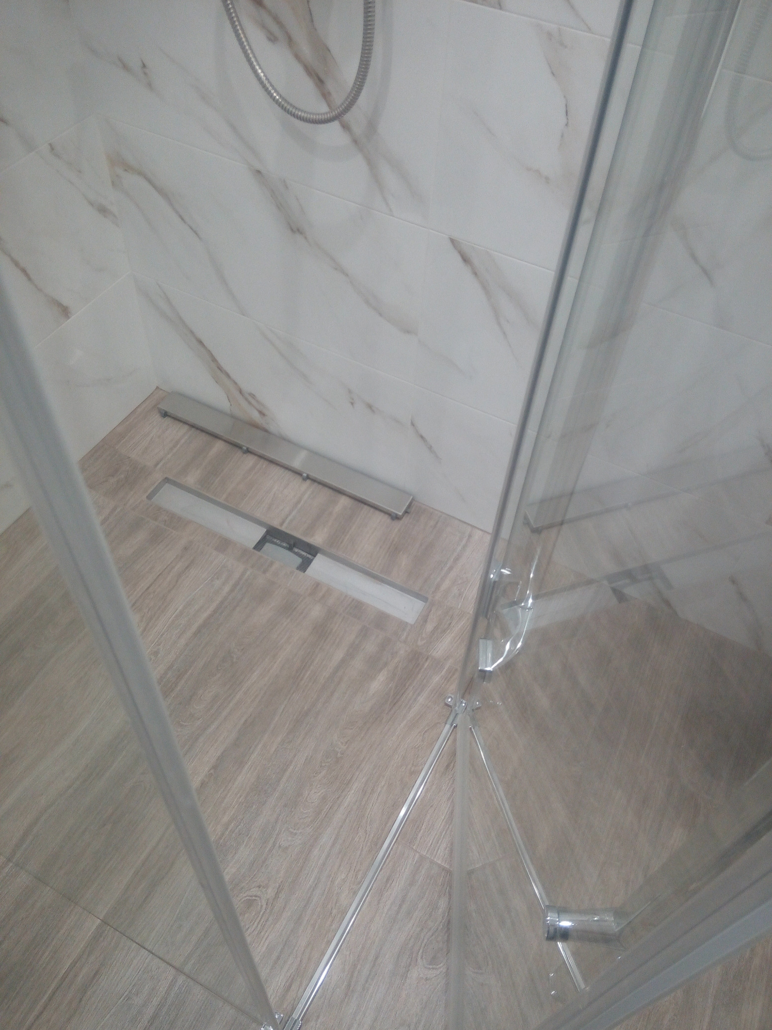 Ukończona kabina prysznicowa z odpływem liniowym w podłodze wyłożonej płytkami imitującymi drewno, ściany wykończone płytkami marmuropodobnymi, widoczny fragment słuchawki prysznicowej.