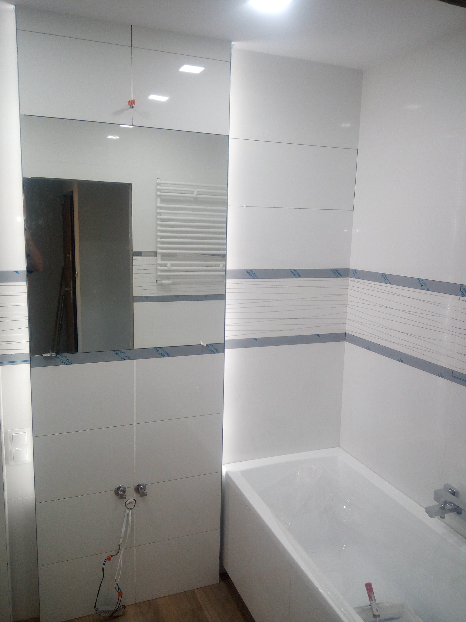 Biała łazienka w trakcie wykańczania, widoczne płytki z folią ochronną, wanna zabezpieczona folią, lustro z odbiciem grzejnika i drabiny, niedokończone podłączenie baterii.