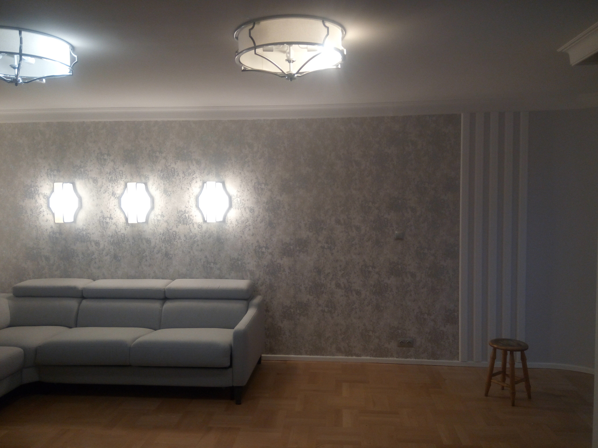 Szary narożnik z podnoszonymi zagłówkami na tle ściany z efektem betonu, oświetlonej trzema kinkietami i dwoma lampami sufitowymi, na parkiecie, z widocznym stołkiem barowym w rogu.