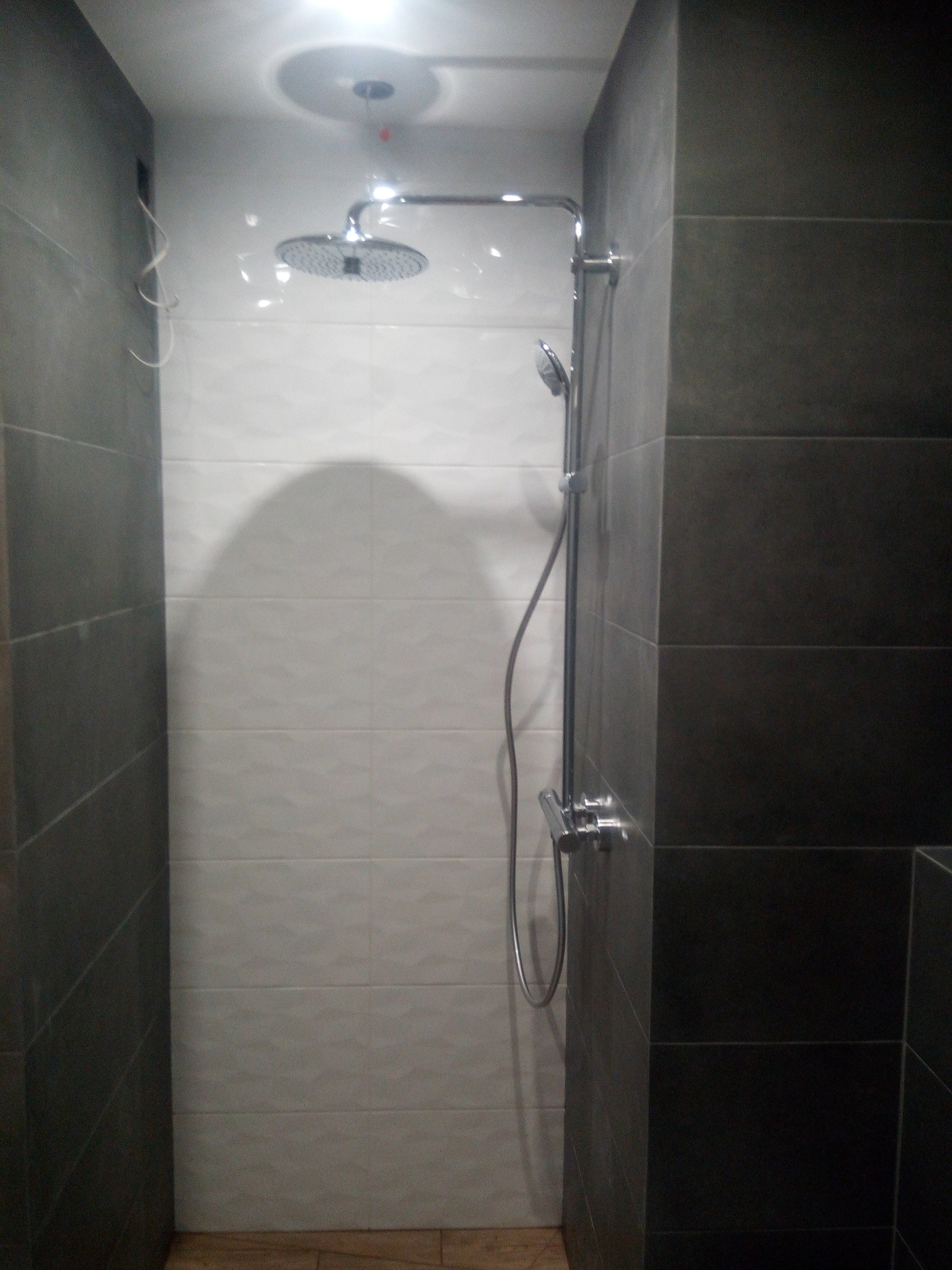 Kabina prysznicowa z deszczownicą i słuchawką, wykończona ciemnymi i jasnymi płytkami, widoczne niedociągnięcia w postaci wystających kabli.