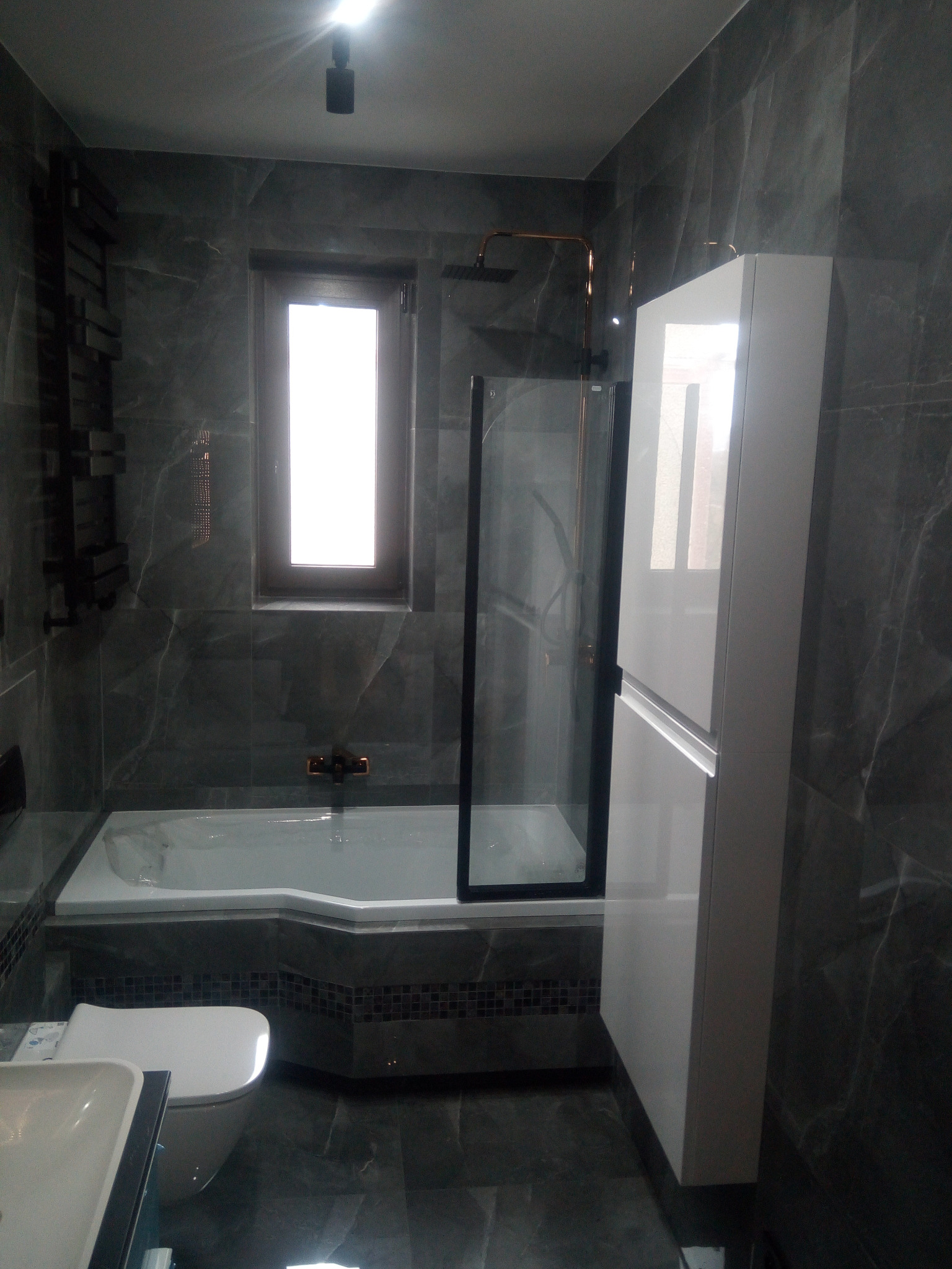 Nowoczesna łazienka wykończona w szarym marmurze z wanną, kabiną prysznicową typu walk-in z czarnymi profilami, podwieszaną szafką i grzejnikiem drabinkowym.