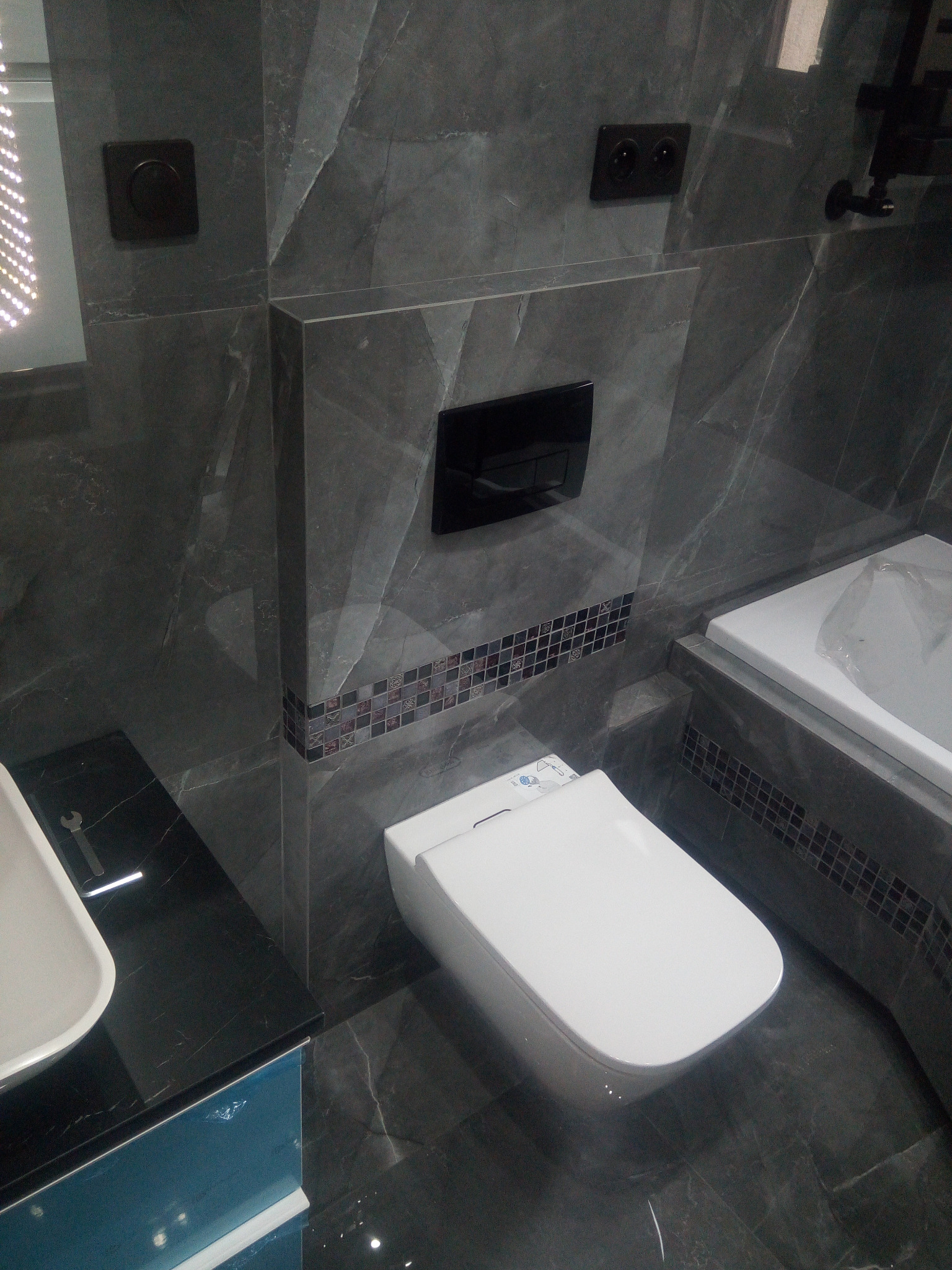 Nowoczesna toaleta myjąca w łazience wykończonej szarym marmurem, z czarnym panelem sterowania i mozaiką dekoracyjną, widoczny fragment umywalki z kluczem płaskim.