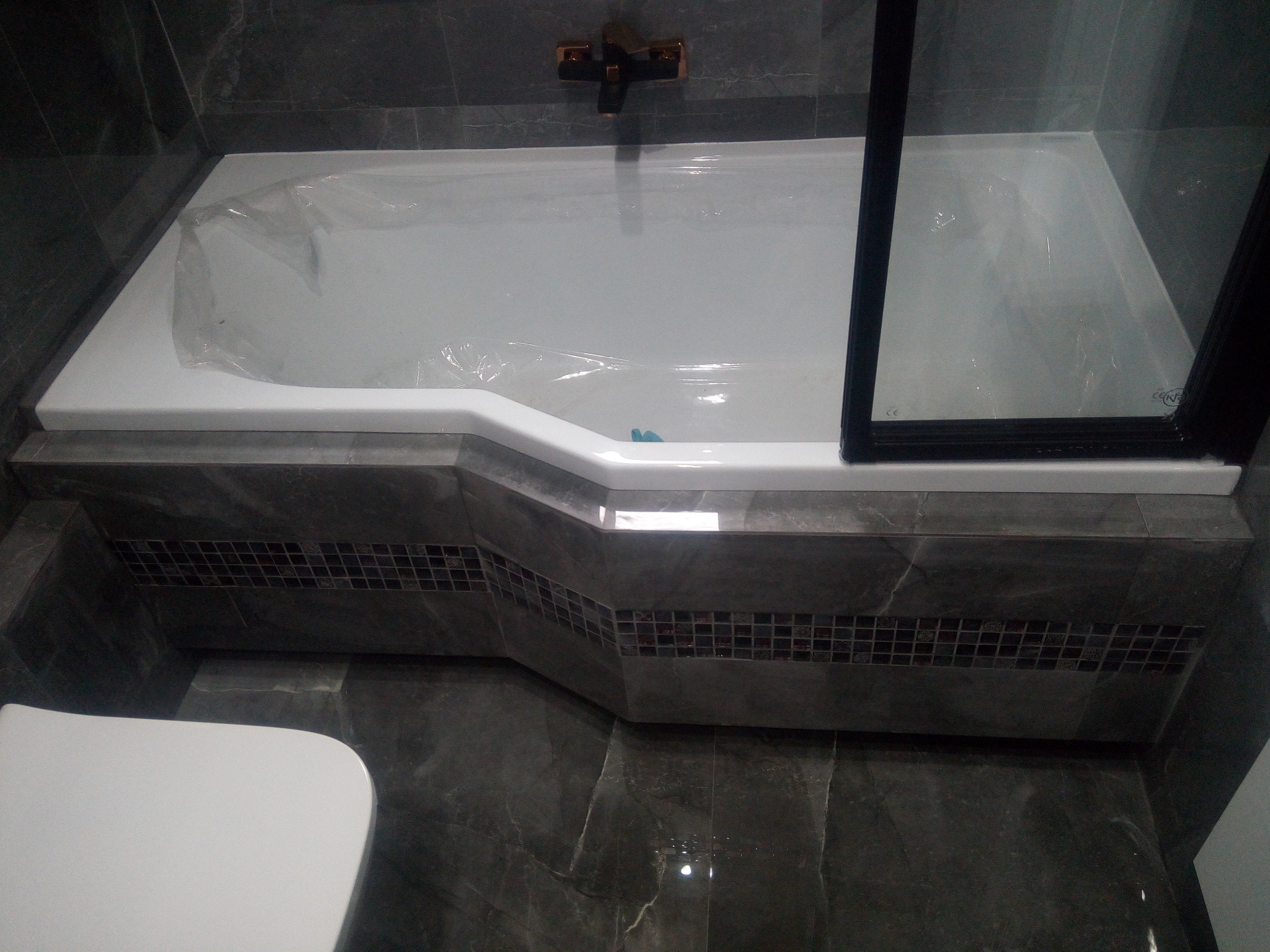 Nowa wanna z czarną armaturą i szklanym parawanem, zabezpieczona folią ochronną, obudowana płytkami z mozaiką, na tle szarych płytek podłogowych i ściennych.