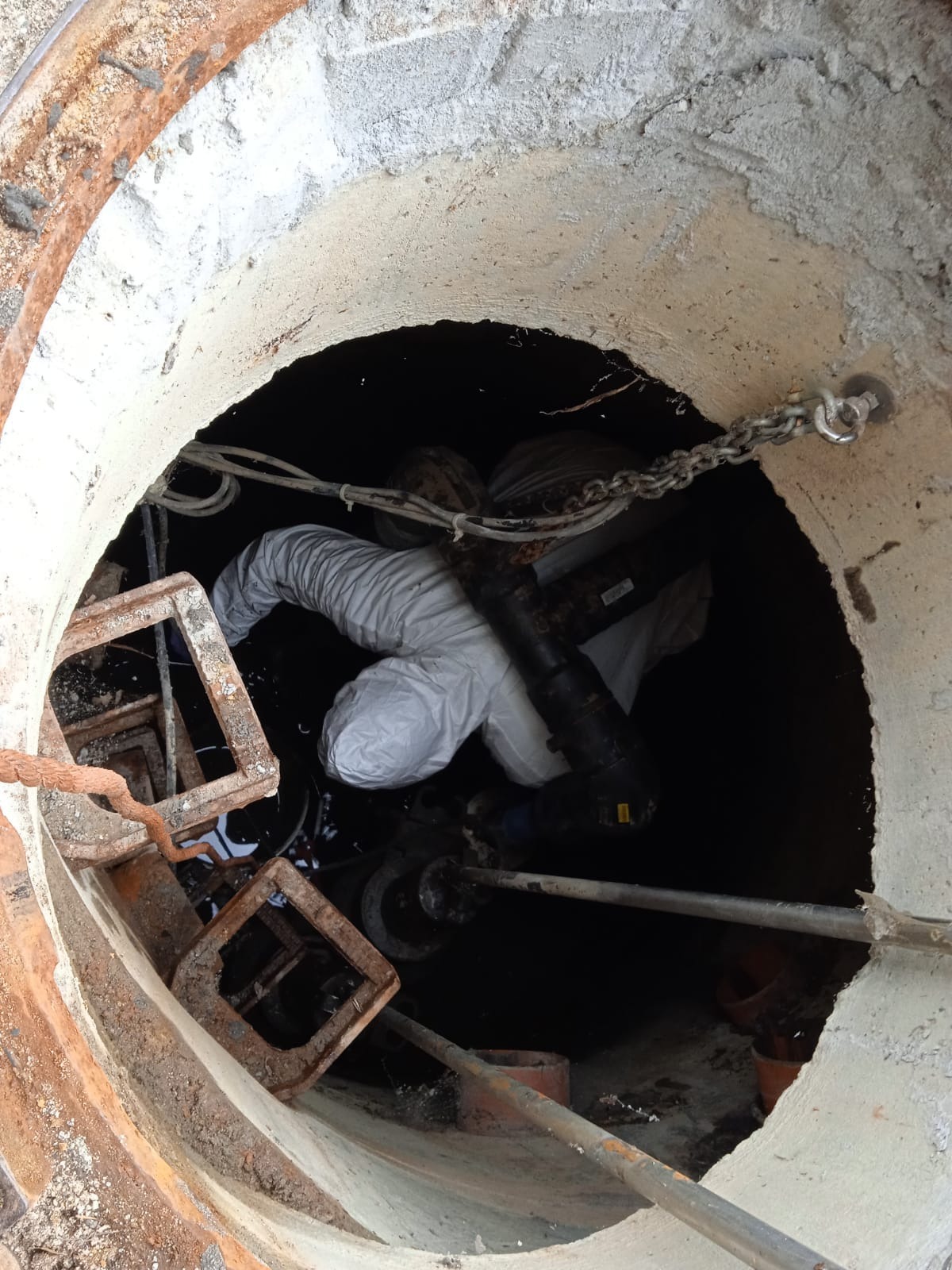 Pracownik w kombinezonie ochronnym, wewnątrz studzienki kanalizacyjnej, używa specjalistycznego sprzętu do czyszczenia lub inspekcji rur.