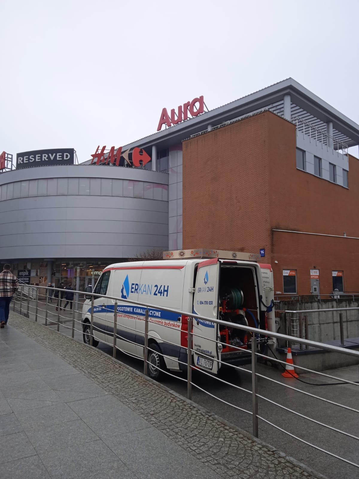 Samochód firmy ERKAN 24H z otwartymi drzwiami bagażnika, z widocznym sprzętem do udrażniania kanalizacji, zaparkowany przed centrum handlowym Aura w pochmurny dzień.