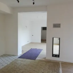 Wnętrze w trakcie remontu: częściowo położona podłoga z wzorzystych płytek, zabezpieczona folią; w przejściu widoczny nowy parkiet zabezpieczony matą; ściany pomalowane na biało, widoczne puszki...