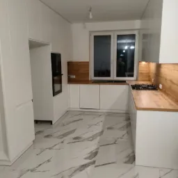 Nowoczesna, biała kuchnia z drewnianymi blatami i marmurową podłogą, widok na okno z drabinką w tle.