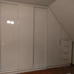 Biała szafa wnękowa z przesuwnymi drzwiami i szufladami, dopasowana do skosu poddasza, widoczna podłoga z paneli.