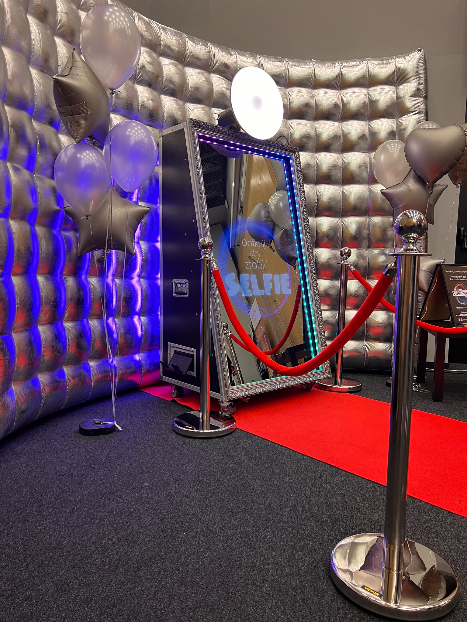 Srebrna ścianka dekoracyjna z balonami i lustrzana konstrukcja z napisem 'Selfie' na czerwonym dywanie, otoczona słupkami z czerwonym sznurem.