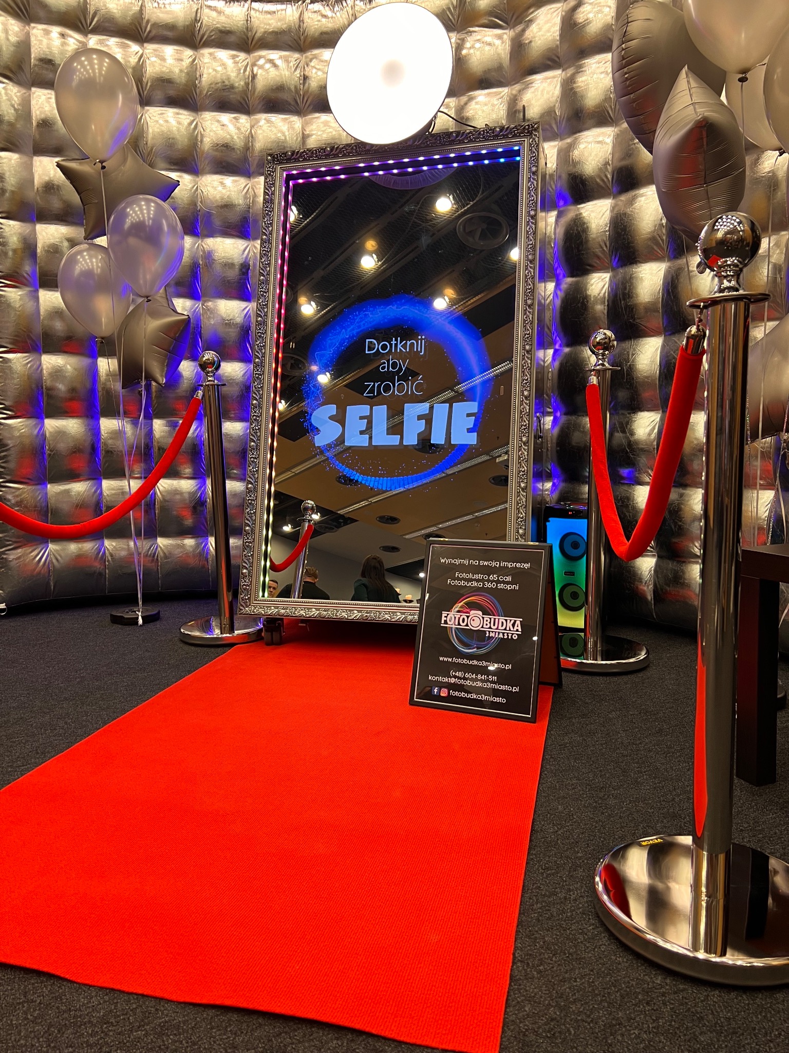 Srebrna ścianka dekoracyjna, balony i czerwony dywan przed lustrem z napisem 'Dotknij aby zrobić SELFIE', otoczonym ledowymi światełkami, obok tabliczka reklamowa.