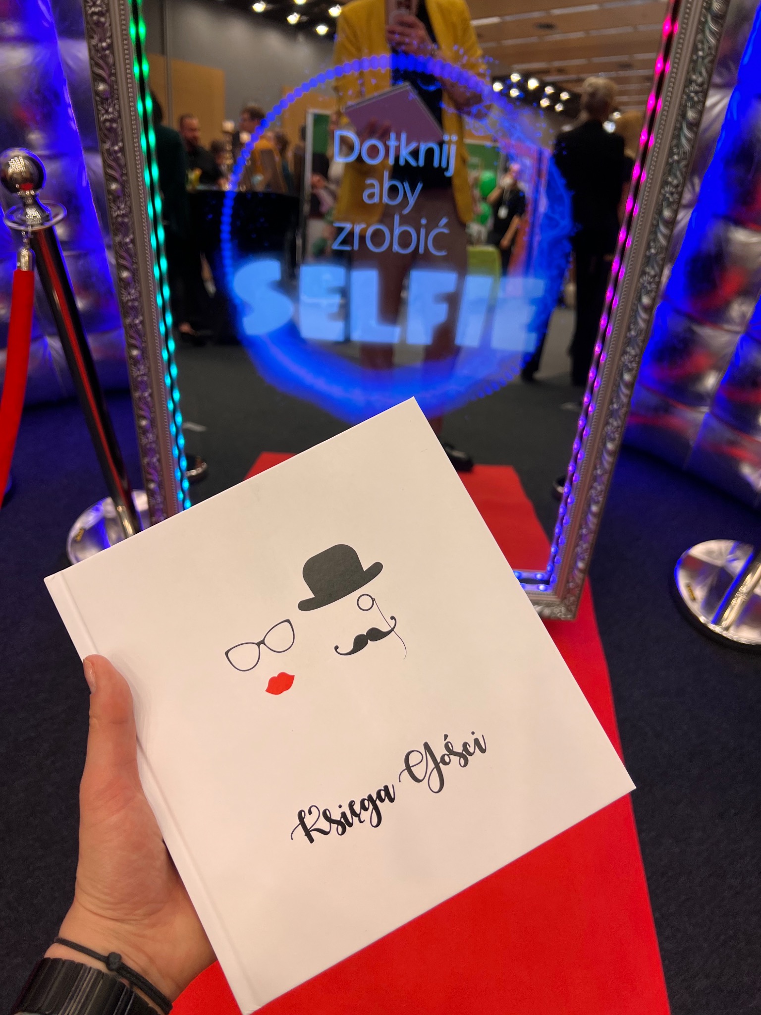 Księga gości z rysunkami wąsów, cylindra i okularów na tle lustra z napisem 'Dotknij aby zrobić SELFIE' i dekoracjami świetlnymi.