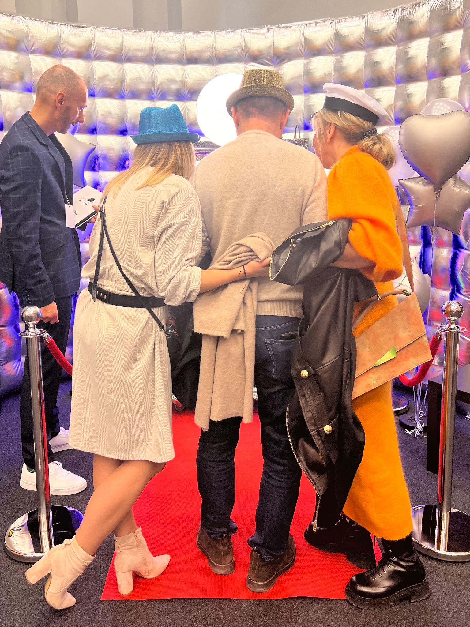 Grupa osób z tyłu na czerwonym dywanie przed srebrnym tłem z balonami w kształcie serc, pozujących do zdjęcia w kapeluszach.