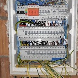 HAEGEN CYPRIAN HAEGENBARTH - Rozdzielnia elektryczna w białej obudowie, z widocznymi przewodami i zabezpieczeniami, zamontowana w ścianie z cegły. Widok z góry.