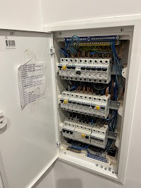 Otwarta skrzynka elektryczna z bezpiecznikami i przewodami na jasnej ścianie, widoczna naklejka z opisami obwodów. Ujęcie z bliska, oświetlenie naturalne.