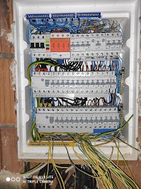 Rozdzielnia elektryczna w białej obudowie, z widocznymi przewodami i zabezpieczeniami, zamontowana w ścianie z cegły. Widok z góry.