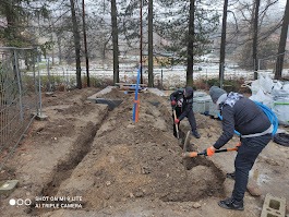 Dwóch mężczyzn kopie rów pod instalację elektryczną na zewnątrz, widoczne rury i narzędzia, teren przygotowany pod budowę, drzewa w tle.