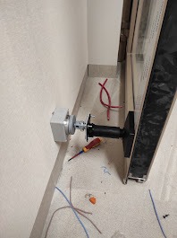 Szara puszka elektryczna zamontowana na ścianie przy drzwiach, z widocznymi przewodami i śrubokrętem. Instalacja w trakcie montażu, surowe wykończenie, jasne tło.