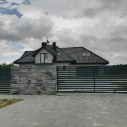 Nowoczesna posesja z przesuwną bramą wjazdową z poziomych profili, betonowe słupki i podmurówka o fakturze kamienia, szara kostka brukowa na podjeździe, dom jednorodzinny w tle.