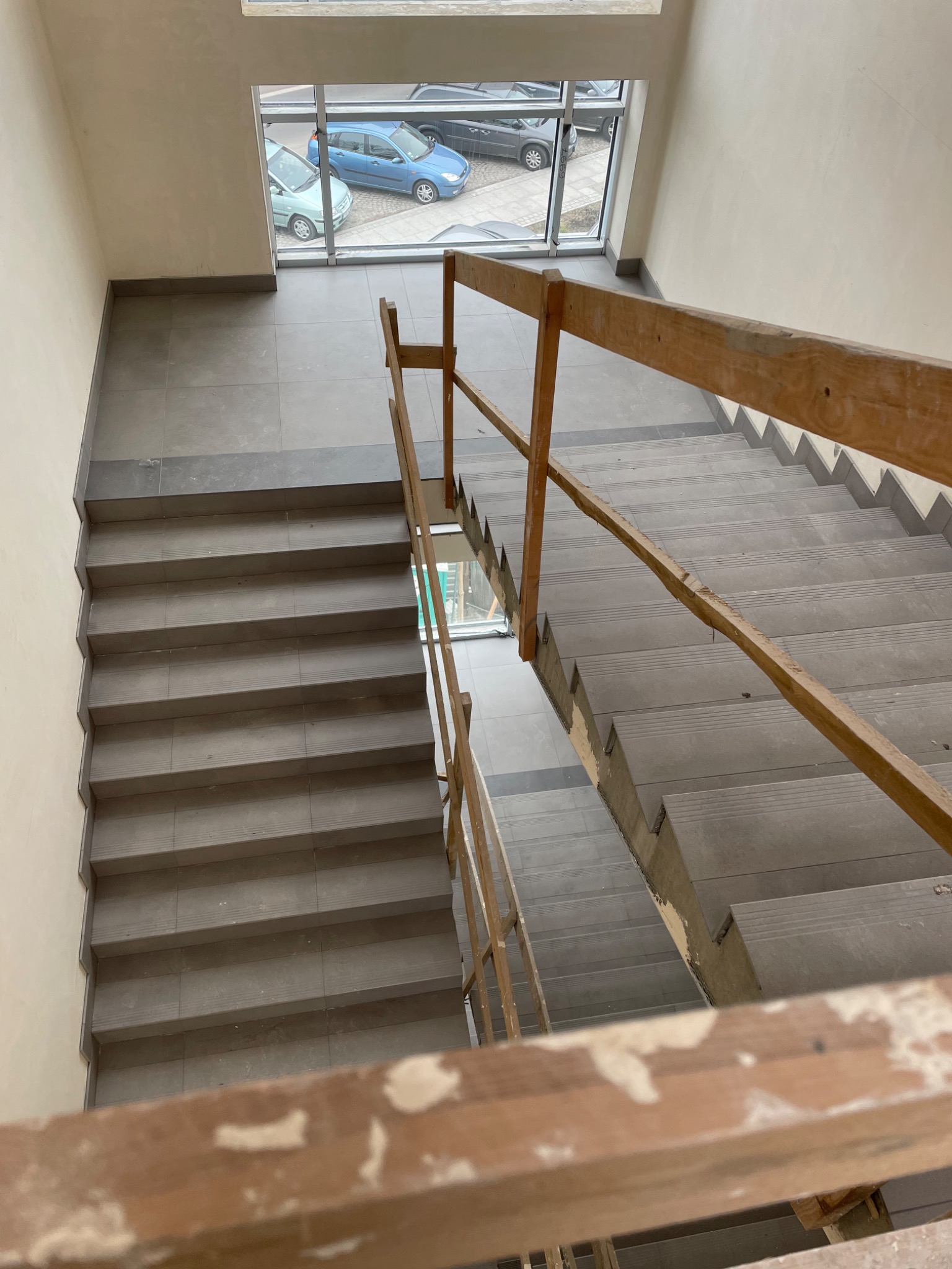 Klatka schodowa w trakcie remontu, widok z góry na schody wyłożone szarą terakotą, prowizoryczna drewniana balustrada z resztkami farby, okno z widokiem na zaparkowane samochody.