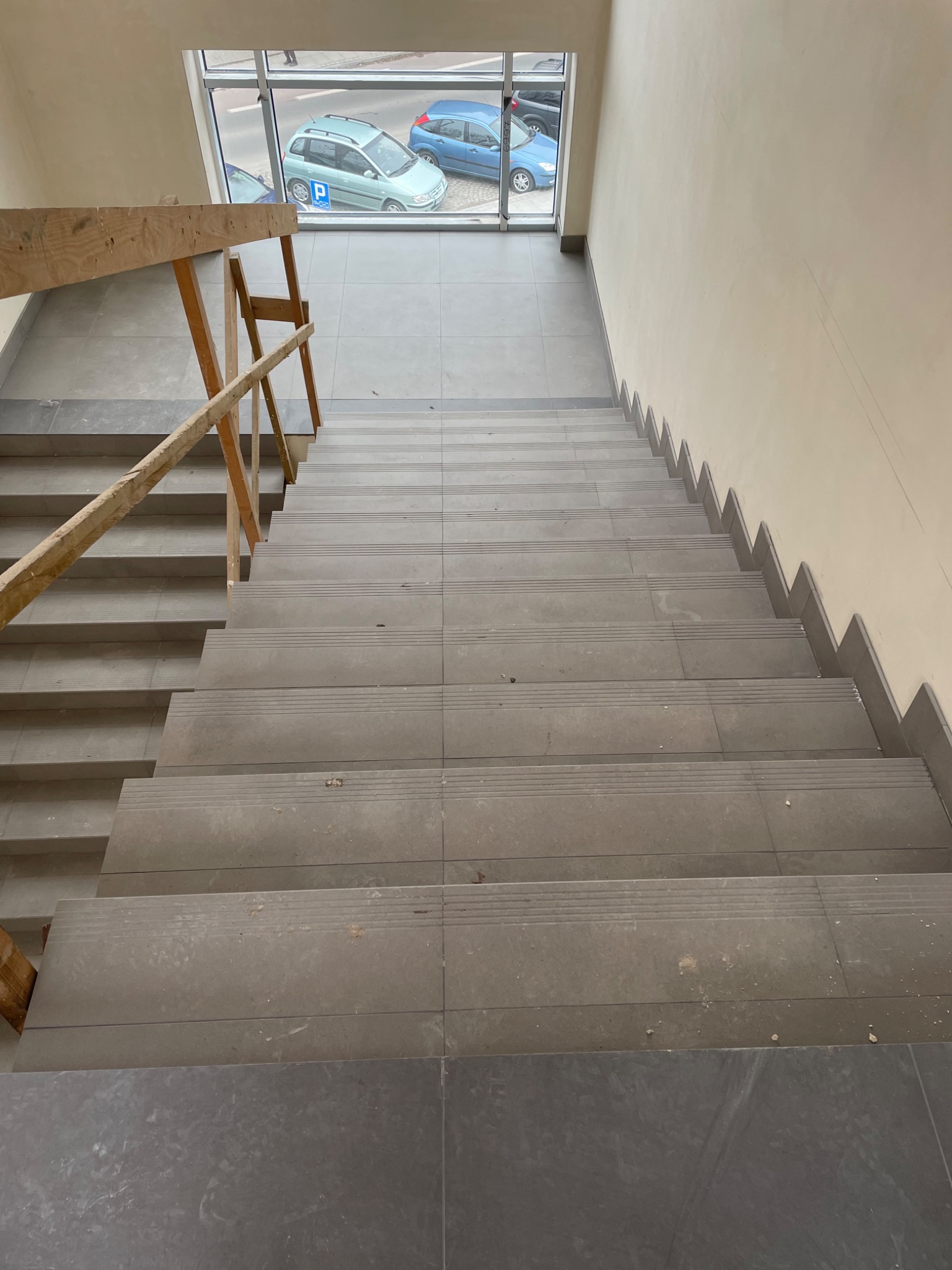 Schody wyłożone szarą płytką ceramiczną z widocznym drewnianym rusztowaniem i oknem na zewnątrz, ukazującym zaparkowane samochody.