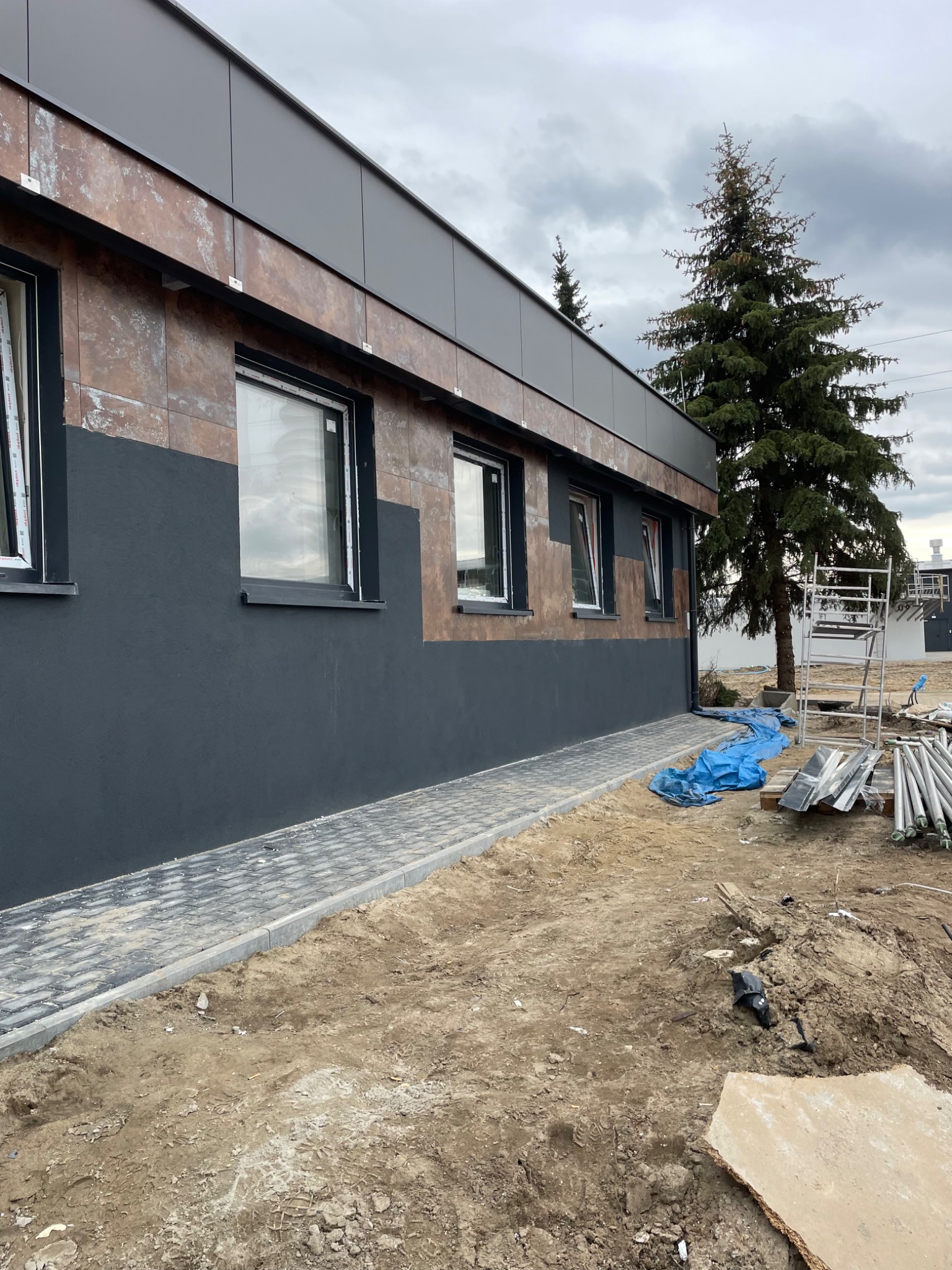 Budynek z częściowo wykończoną elewacją: dolna część szara, górna z brązowych płyt imitujących rdzę, widoczne okna z białymi ramami, obok budynku rusztowanie i materiały budowlane, w tle drzewo...