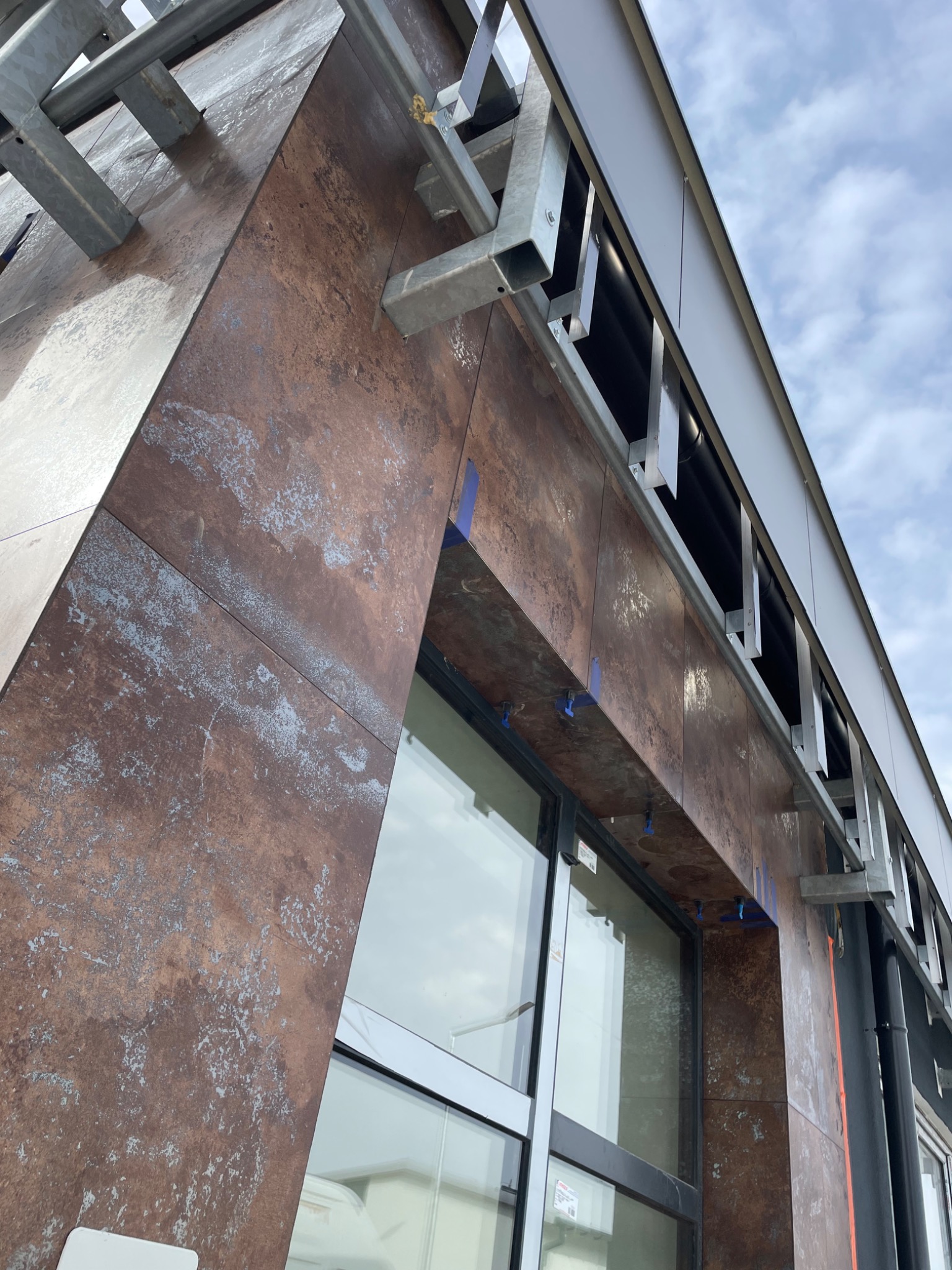 Montaż elewacji z płytek imitujących rdzę, widoczne metalowe elementy konstrukcyjne i niebieskie kliny dystansowe.