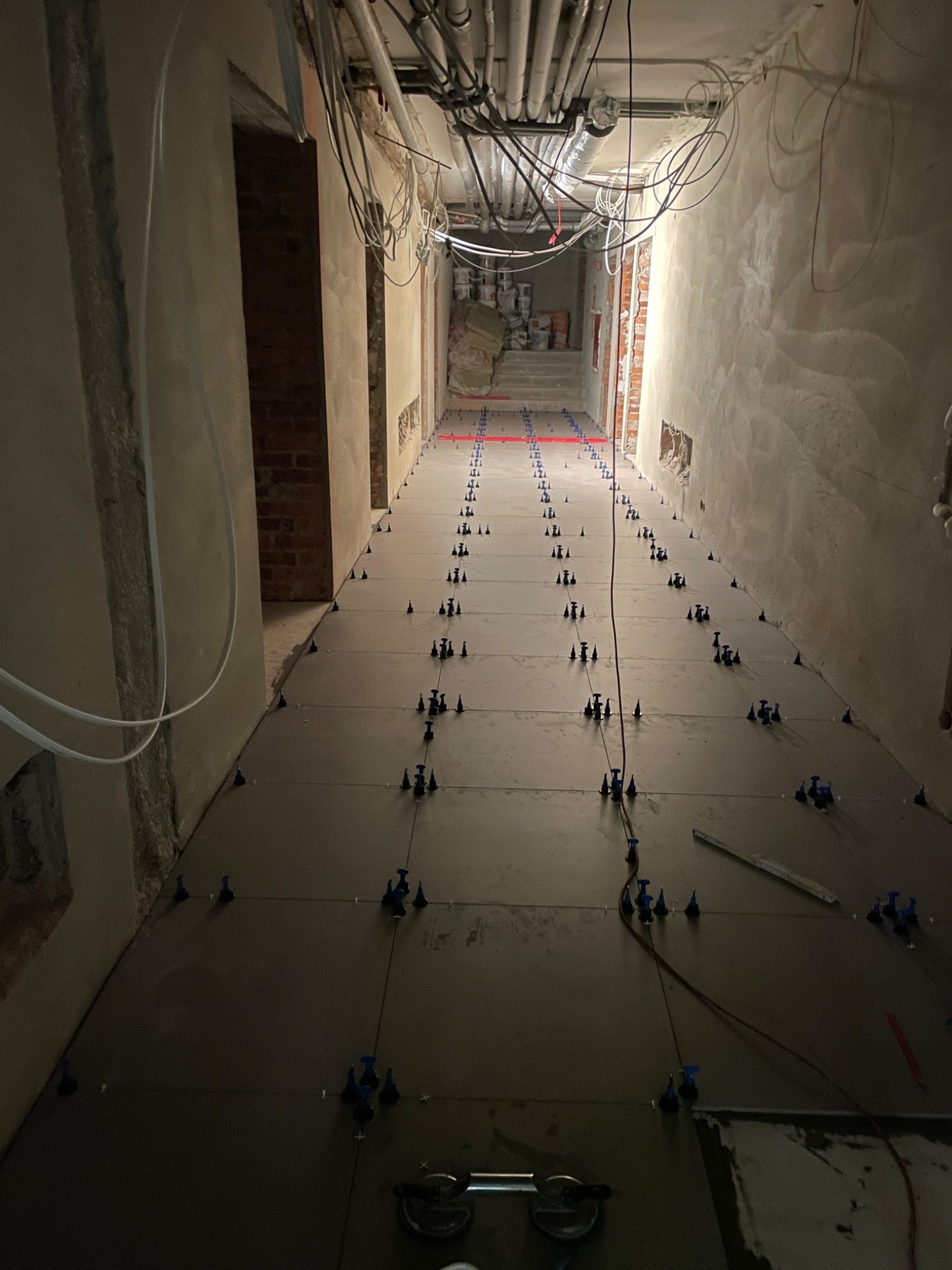 Układanie ciemnych płytek podłogowych w długim korytarzu, widoczne narzędzia glazurnicze i system poziomowania płytek.