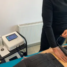 Pacjentka leżąca na turkusowym łóżku podczas zabiegu z użyciem aparatu do terapii, obsługiwanego przez osobę w czarnym stroju, widoczny fragment pleców pacjentki, urządzenie do terapii na wózku...