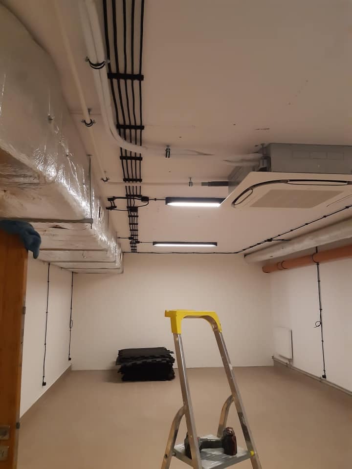Piwnica po remoncie z widocznymi przewodami elektrycznymi ułożonymi na suficie, klimatyzacją i drabiną z żółtymi elementami.