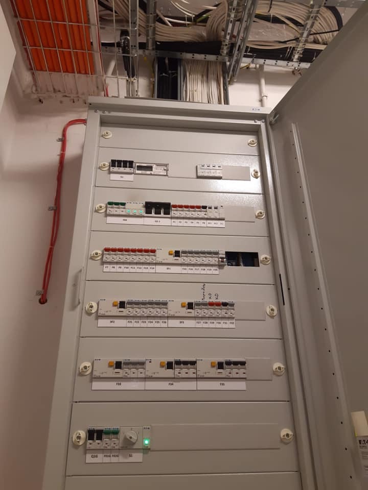 Otwarta szafa sterownicza z widocznymi zabezpieczeniami i przewodami w korytkach kablowych pod sufitem.