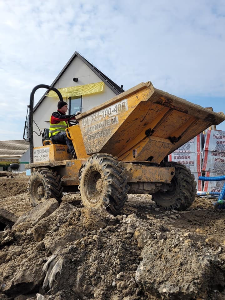 Żółta wywrotka budowlana z numerem telefonu na korpusie, prowadzona przez operatora w kamizelce odblaskowej, na tle budowy domu z zabezpieczoną folią elewacją.