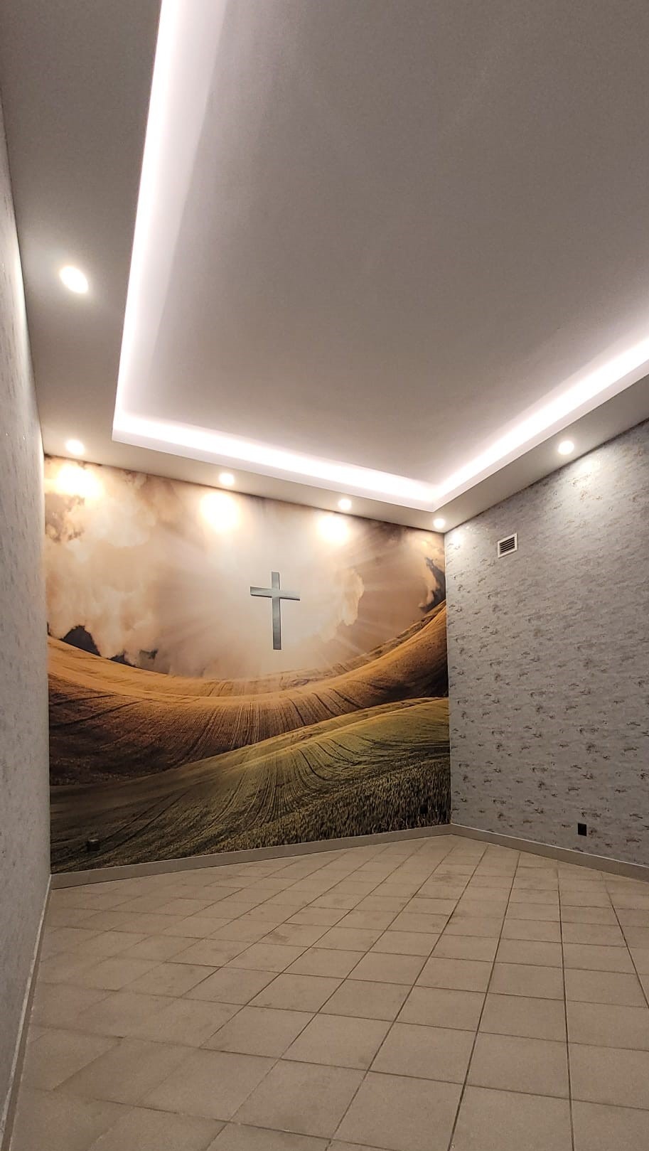 Wnętrze pokoju z podświetlanym sufitem, tapetą przedstawiającą krajobraz z krzyżem i płytkami na podłodze.