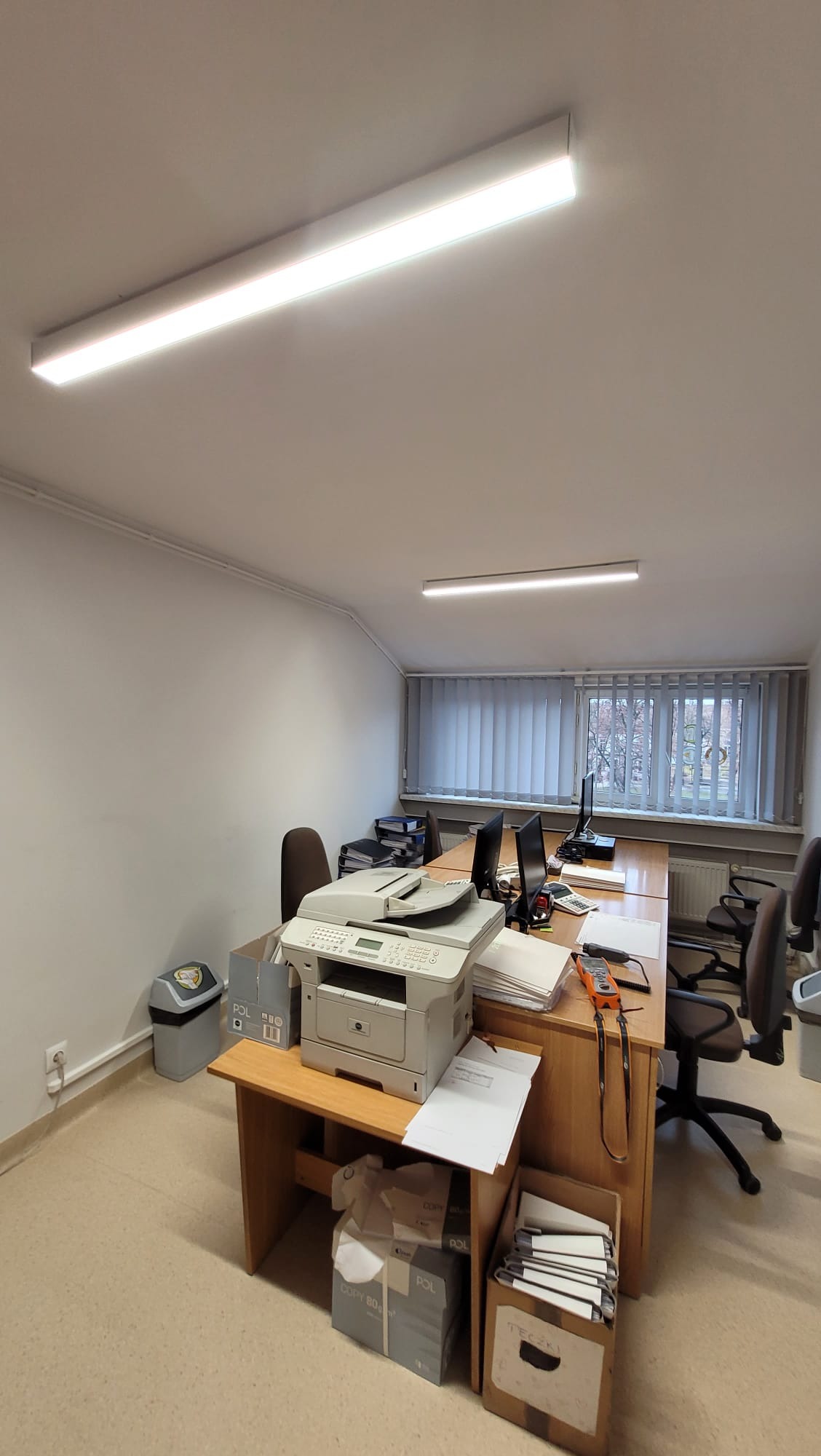 Biuro z dwoma biurkami, krzesłami, komputerami i drukarką. Widoczne podwieszane lampy jarzeniowe oświetlające pomieszczenie. Na biurku leżą papiery i miernik elektryczny.