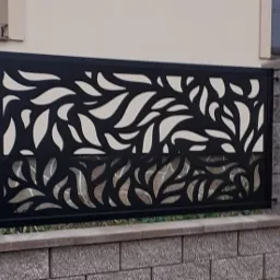 Czarna, metalowa osłona z wycinanym wzorem liści, zamontowana na murku z szarej cegły przy budynku.