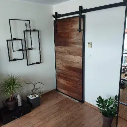 Drewniane drzwi przesuwne w stylu rustykalnym z czarnymi elementami metalowymi, zamontowane na szynie, w jasnym wnętrzu z drewnianą podłogą, dekoracjami ściennymi i roślinami doniczkowymi.