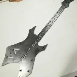 Metalowa gitara z wyciętym napisem 'FILIP SZUBA RECORDINGS' leżąca na białej powierzchni.