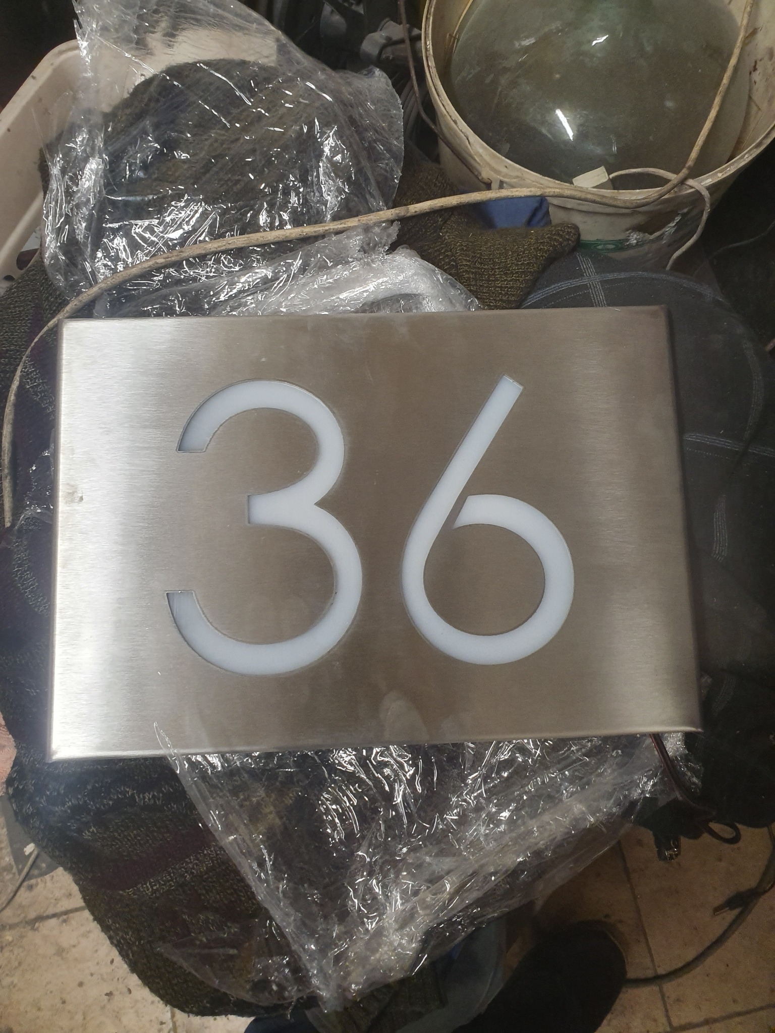 Błyszcząca, prostokątna tabliczka z numerem '36' wyciętym laserowo, podświetlonym od spodu białym światłem LED, leżąca na tle roboczym z folią i innymi przedmiotami.