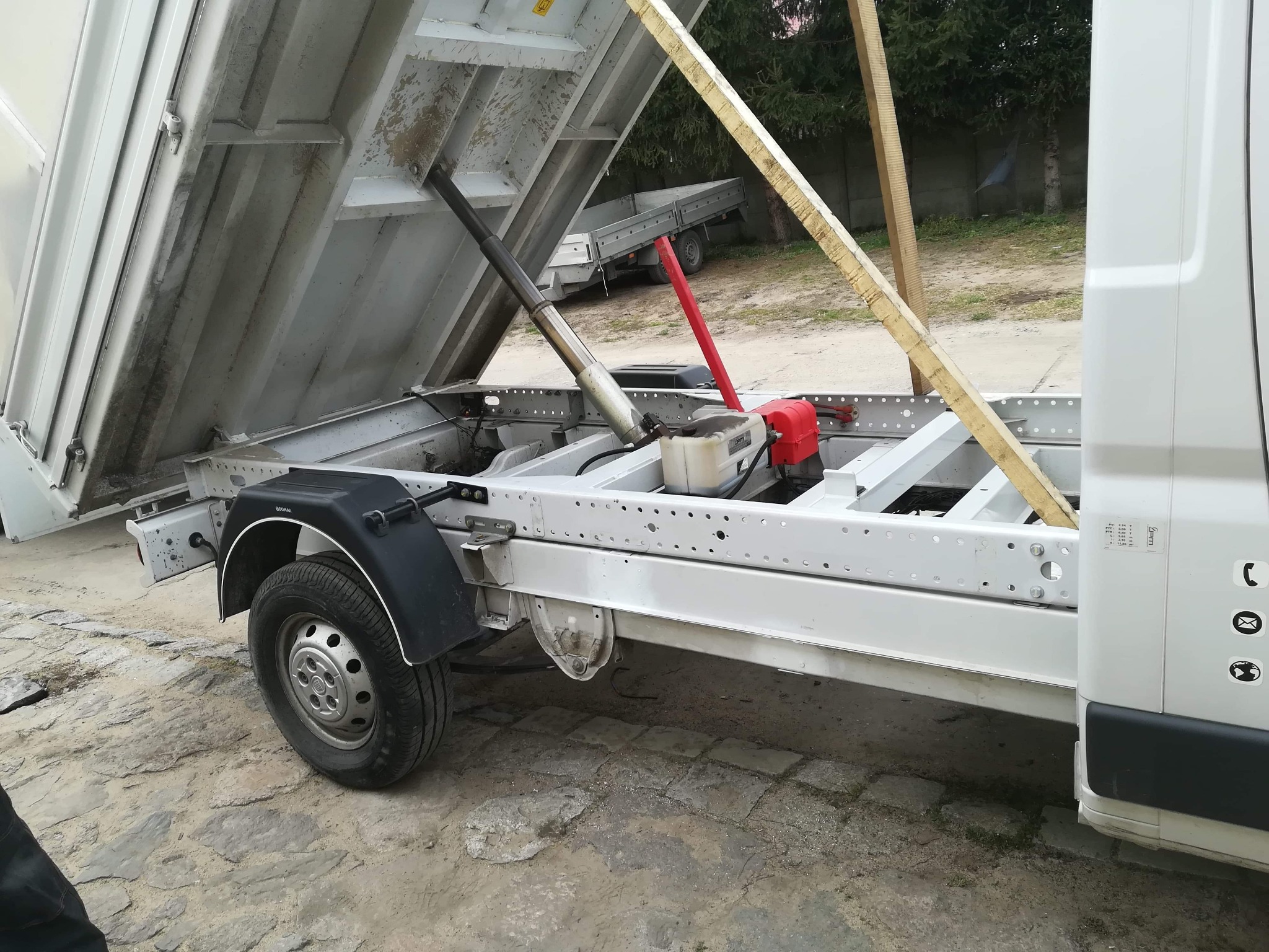 Ciężarówka z podniesioną skrzynią ładunkową, widoczny mechanizm podnoszenia z siłownikiem hydraulicznym i drewniane podpory, w tle drugi pojazd i drzewa.