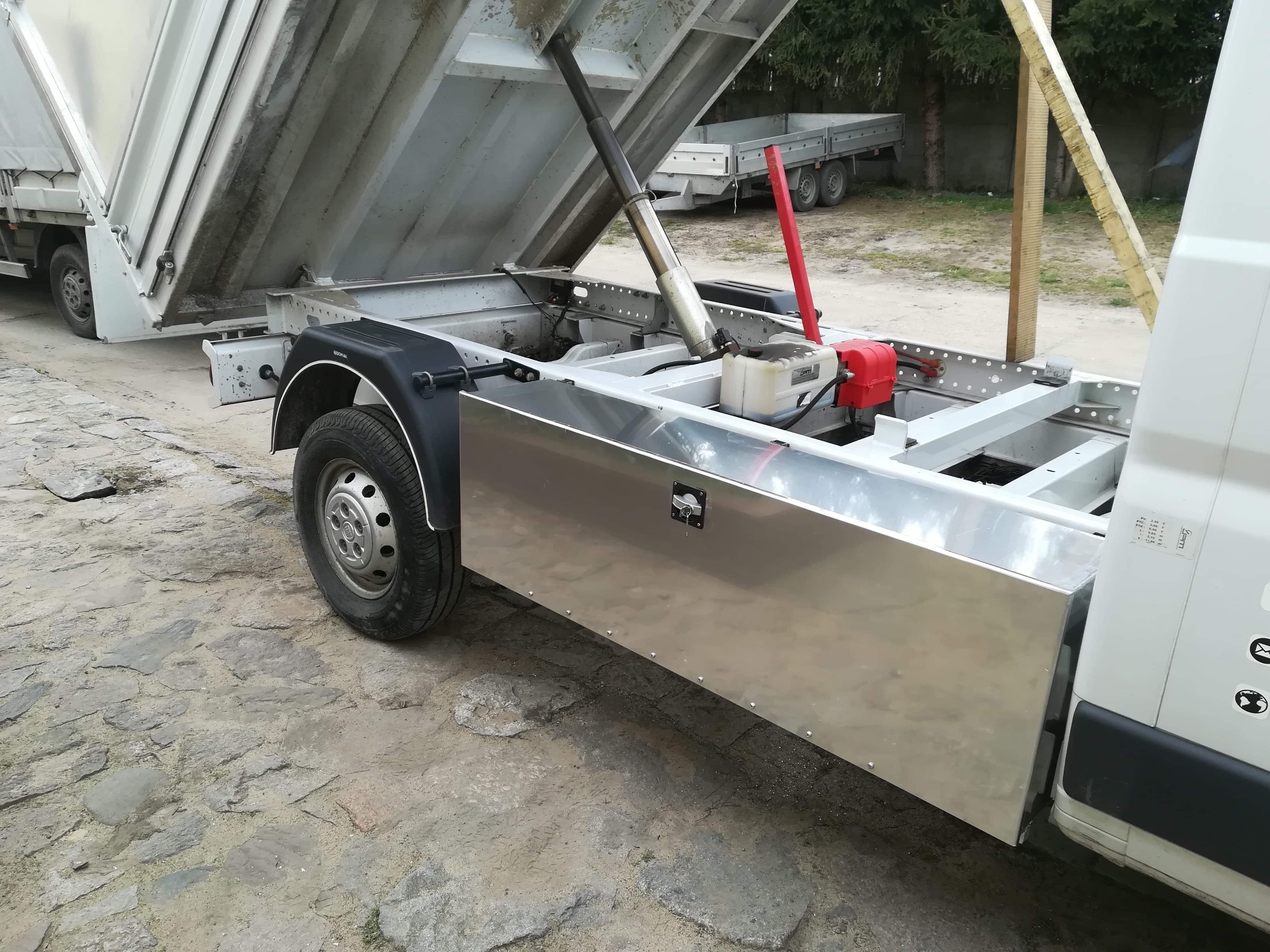 Podniesiona skrzynia ładunkowa wywrotki z widocznym siłownikiem hydraulicznym, rama nośna i aluminiowe panele boczne, na nierównym podłożu z kostki.